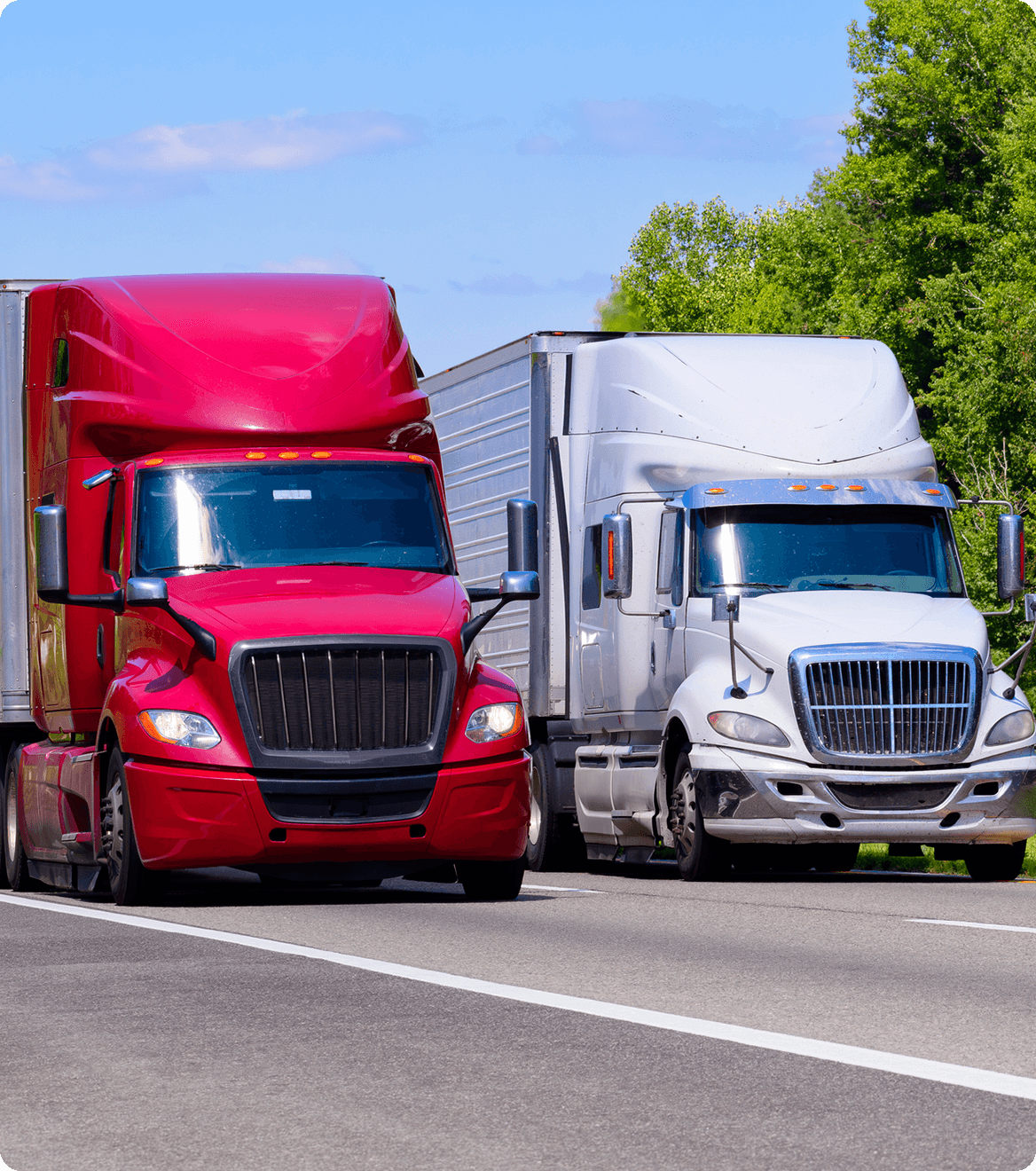 Two semi-trucks, one red and one white, driving side by side on a highway, with trees and a clear sky in the background.