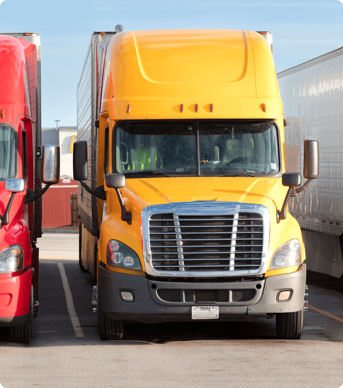 Yellow semi-truck parked between a red truck and a white trailer in a parking lot under a clear blue sky.