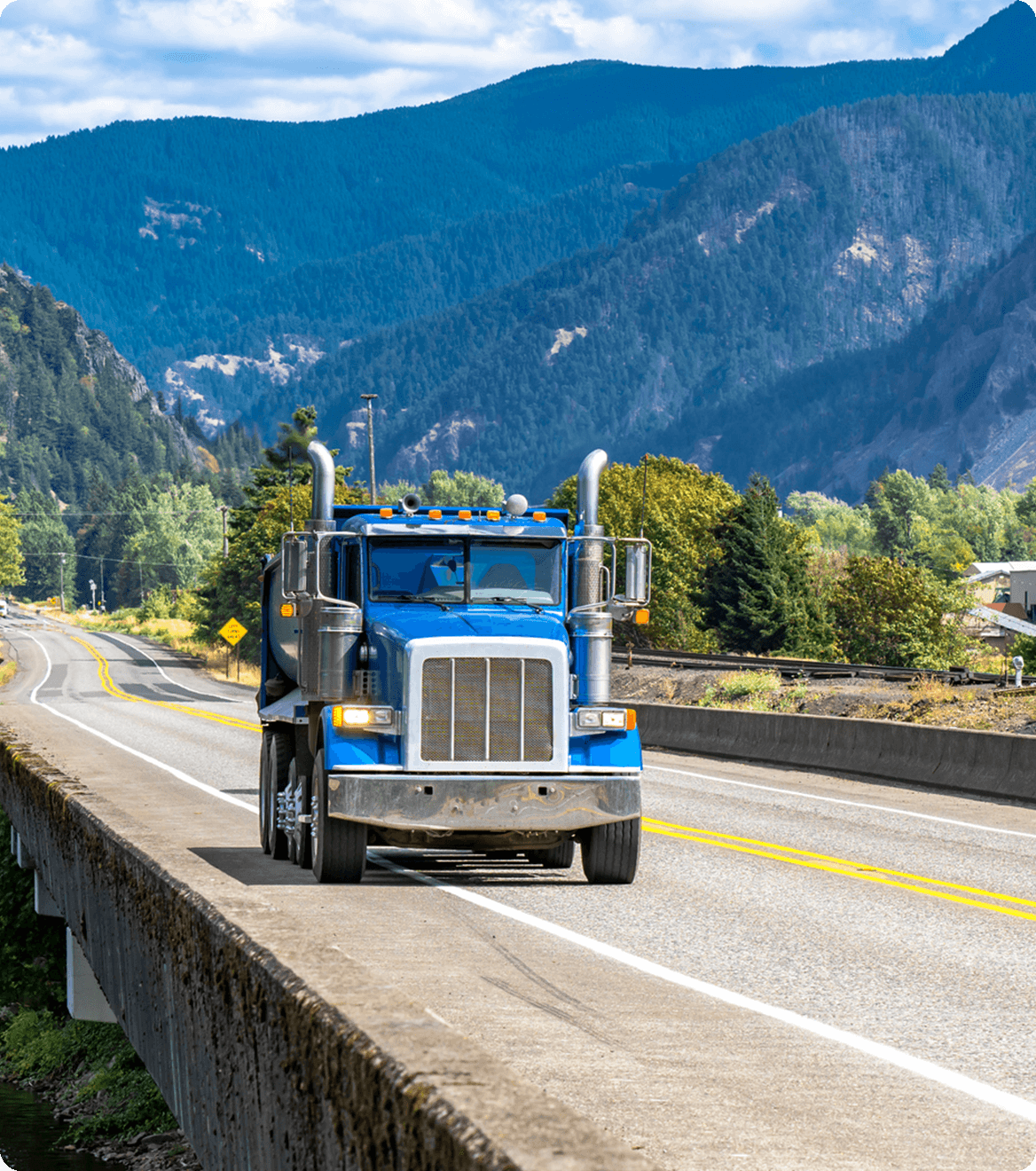 Blue semi-truck driving on a mountain road with trees and mountains in the background under a partly cloudy sky.