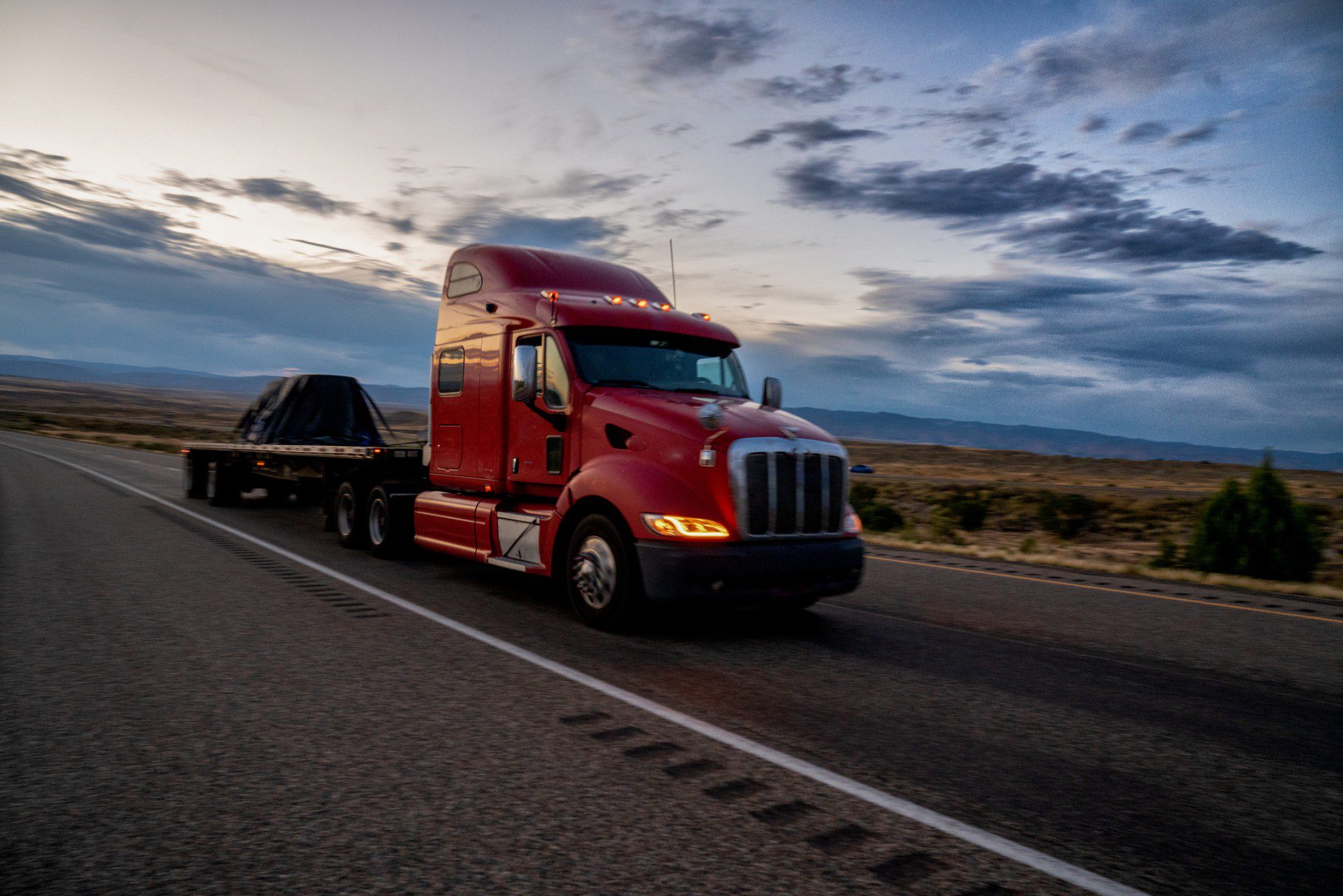 A red semi-truck with a flatbed trailer drives along a highway at dusk, with a cloudy sky and open landscape in the background.